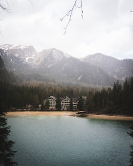 A serene mountain landscape featuring a calm lake in the foreground. The shoreline has a sandy beach with several large, chalet-style buildings nestled among dense coniferous forest. Snow-capped mountains rise majestically in the background, and branches are visible at the top of the image, framing the scene.