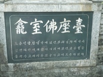 A stone plaque featuring Korean text inscribed on a dark background, bordered by a light gray stone frame. The text is large and prominent at the top, with smaller characters below.