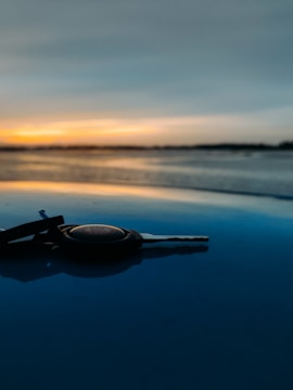 A car key rests on a smooth, shiny surface with a serene sunset in the background. The sky features a gradient of colors ranging from orange to blue, reflecting softly on the surface.