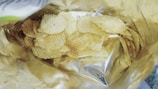 A partially opened bag contains a pile of ridged potato chips. The bag has a metallic interior, reflecting light, and the chips are lightly seasoned with visible specks.
