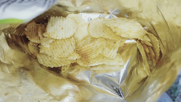 A partially opened bag contains a pile of ridged potato chips. The bag has a metallic interior, reflecting light, and the chips are lightly seasoned with visible specks.