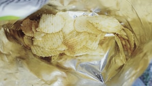 A partially opened bag contains a pile of ridged potato chips. The bag has a metallic interior, reflecting light, and the chips are lightly seasoned with visible specks.