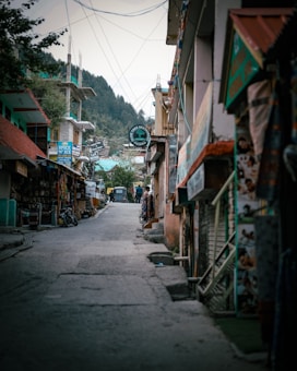 A narrow street lined with small shops on either side, featuring colorful signs and banners. The road is slightly inclined, leading towards a backdrop of lush green hills. A few people are seen walking down the street, and a vehicle is parked at the far end. The atmosphere appears to be relaxed and somewhat remote.