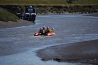 A motorized inflatable boat with four people wearing helmets and life jackets is navigating a narrow, calm river with muddy banks. In the background, a small, stationary boat is moored to the riverbank.