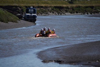 A motorized inflatable boat with four people wearing helmets and life jackets is navigating a narrow, calm river with muddy banks. In the background, a small, stationary boat is moored to the riverbank.