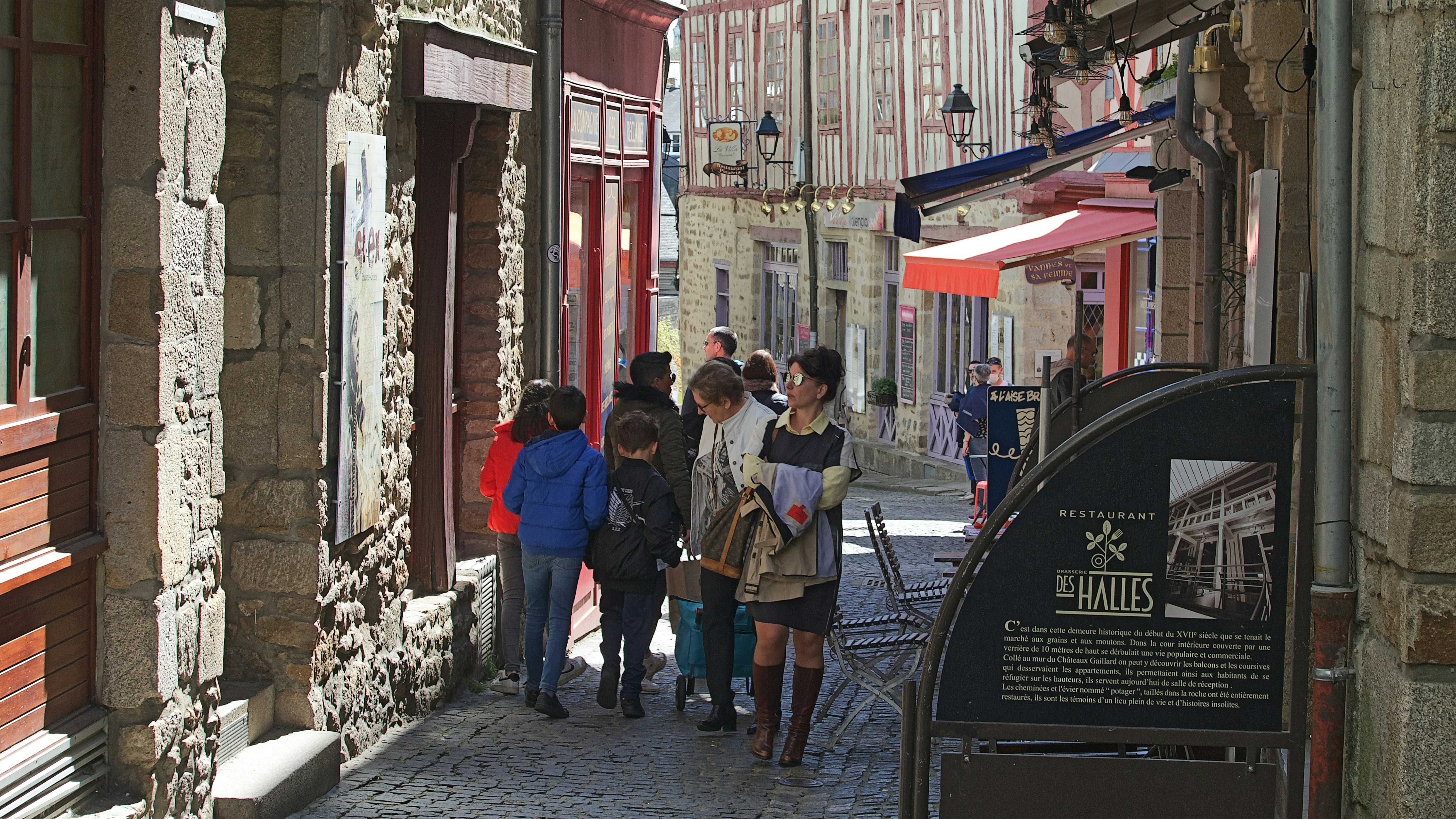 People walk along a narrow cobblestone street in Dinan, France, lined with restaurants and half-timbered buildings.