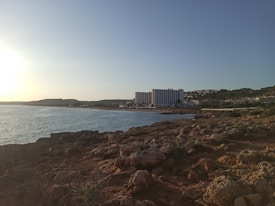 A rocky coastline with a large hotel in the background. The sun is setting on the horizon, casting a warm glow over the water and the surrounding landscape. The area is mostly undeveloped except for the hotel, with a natural and rugged terrain leading up to the water's edge.