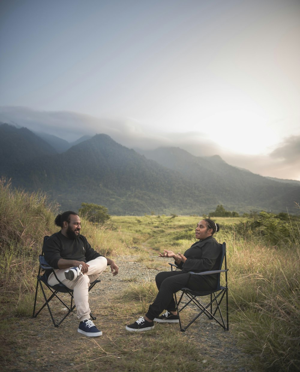 a man and woman sitting in chairs in a grassy field with mountains in the background