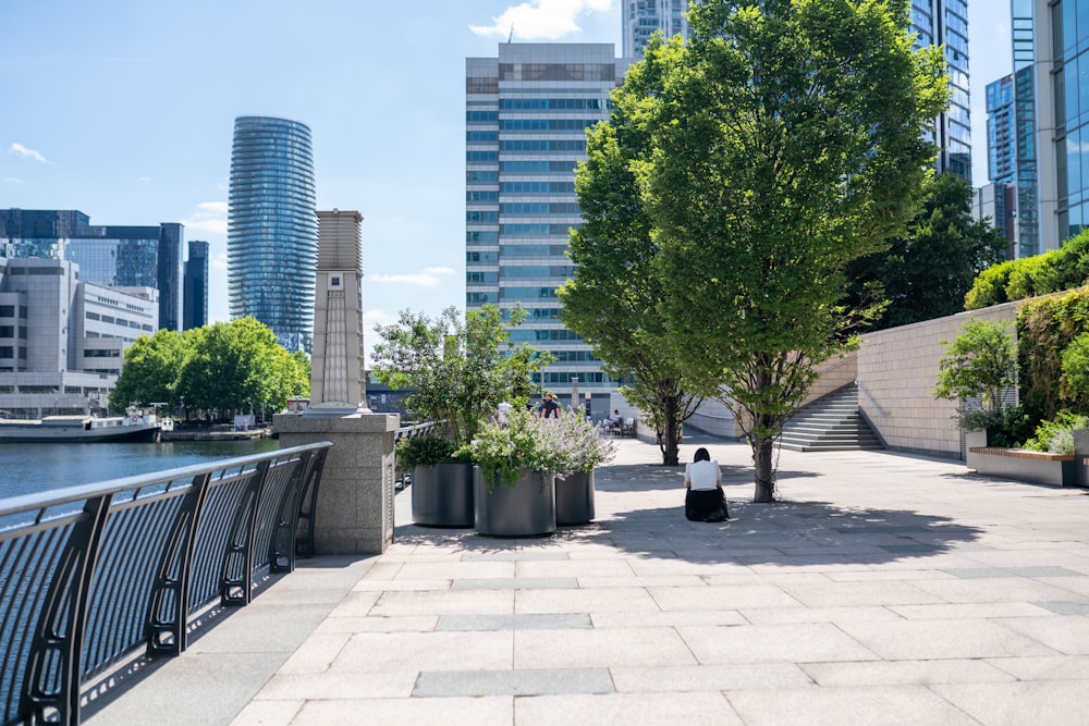 a person sitting on a stone walkway by a body of water with tall buildings in the background