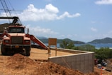 A construction site with a red dump truck parked on dirt terrain. In the background, there are cranes and unfinished concrete structures. The landscape view includes mountains, a body of water, and a partly cloudy sky.