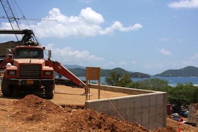 A construction site with a red dump truck parked on dirt terrain. In the background, there are cranes and unfinished concrete structures. The landscape view includes mountains, a body of water, and a partly cloudy sky.