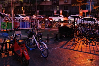 Bicycles and scooters are parked in front of a fence, while cars are on the street in the background. The ground is wet, possibly due to recent rain, and the area is illuminated by streetlights casting warm, orange tones. Shadows from the railing create interesting patterns on the wet surface.