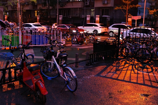 Bicycles and scooters are parked in front of a fence, while cars are on the street in the background. The ground is wet, possibly due to recent rain, and the area is illuminated by streetlights casting warm, orange tones. Shadows from the railing create interesting patterns on the wet surface.