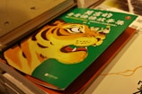 A vibrant illustration features the head of a tiger on a green book cover. The tiger's expression is fierce, with visible teeth and expressive eyes. In the background, there's a small cartoon depiction of people in traditional attire. The book cover has text in Chinese characters, suggesting it might be a collection of stories or an educational book.