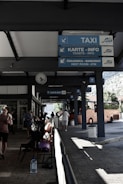 A bus station with signs indicating services such as taxi, tickets, restroom, and ATM. People are sitting on benches, with some holding luggage. A bright orange bus is parked nearby. The station is partially shaded, with sunlight creating patterns on the ground.