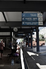 A bus station with signs indicating services such as taxi, tickets, restroom, and ATM. People are sitting on benches, with some holding luggage. A bright orange bus is parked nearby. The station is partially shaded, with sunlight creating patterns on the ground.