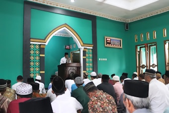 A group of men is seated in a room with green walls, listening to a speaker dressed in white who stands behind a podium adorned with an ornate arch. The men are wearing different types of headgear, including caps and turbans. The room features an intricate design on the walls and a large digital clock near the ceiling. Light streams in through multiple windows with wood framing on one side of the room.