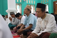 A group of men is seated on a patterned carpet inside a room with teal walls and several windows. They are dressed in traditional clothing and hats, appearing to be engaged in prayer or meditation. The atmosphere is calm and focused.