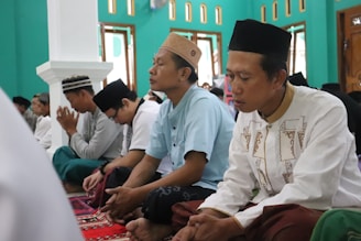 A group of men is seated on a patterned carpet inside a room with teal walls and several windows. They are dressed in traditional clothing and hats, appearing to be engaged in prayer or meditation. The atmosphere is calm and focused.