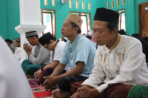 A group of men is seated on a patterned carpet inside a room with teal walls and several windows. They are dressed in traditional clothing and hats, appearing to be engaged in prayer or meditation. The atmosphere is calm and focused.
