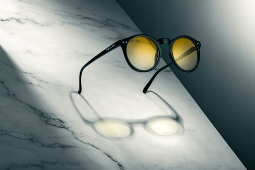 Round sunglasses with yellow-tinted lenses rest on a marble surface, casting a distinct shadow due to the angled lighting. The overall composition emphasizes the sleek design of the eyewear.