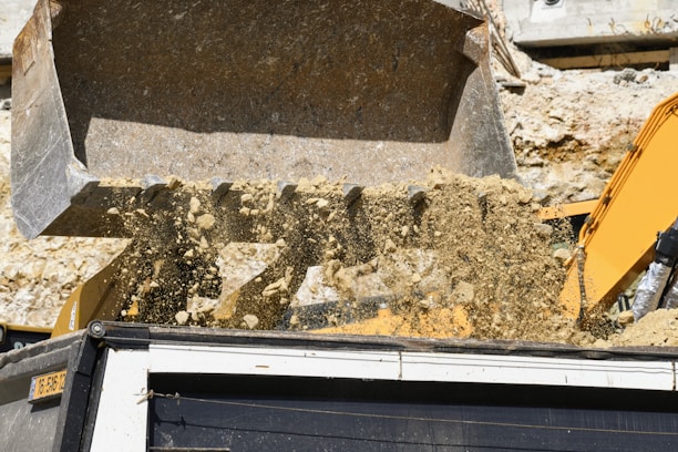A construction vehicle, likely an excavator, is dumping a large load of dirt and debris into a truck. The backdrop shows a rocky or dirt-packed environment, indicative of a construction site or quarry.