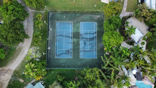An aerial view of a tennis court surrounded by lush green trees and vegetation. The court appears to be outdoors with two separate playing areas marked clearly with white lines. Nearby pathways and sections of a garden or park can be seen, along with a few small structures.