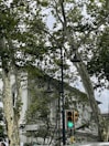 A street scene featuring a classical white building partially covered by large, leafy trees. There is a pedestrian traffic light showing green and a street lamp with two lanterns. A directional sign points to Beşiktaş.