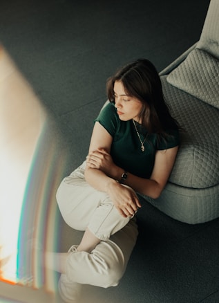 A woman sits on the floor leaning against a cushioned chair. She is wearing a dark green t-shirt and light beige pants. Her hands are gently clasped together on her lap, and she gazes contemplatively to the side. The room is softly lit, creating a calm and introspective atmosphere.