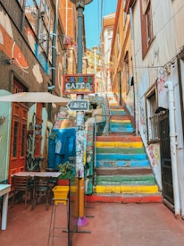 A vibrant alleyway features a set of colorful steps leading upwards, surrounded by buildings adorned with street art and graffiti. Outside seating is visible near a café sign that reads 'Crysol Café,' with another sign pointing to 'Babel.' A fish mural and various stickers and small street decorations are also present, creating a lively and artistic atmosphere.