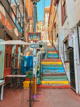 A vibrant alleyway features a set of colorful steps leading upwards, surrounded by buildings adorned with street art and graffiti. Outside seating is visible near a caf&eacute; sign that reads 'Crysol Caf&eacute;,' with another sign pointing to 'Babel.' A fish mural and various stickers and small street decorations are also present, creating a lively and artistic atmosphere.