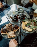 A glass table set with an assortment of brunch items including a plate with waffles topped with chocolate and banana slices, alongside a bowl of chocolate ice cream. Next to it, another plate holds a salad with greens, slices of fruit, and berries. There is a metal tray with two teapots, several glasses, and silverware. Two people are seated at the table, with only their hands visible.