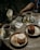A wooden table is set with two plates of desserts, one with a chocolate cake and the other with a vanilla cake. Two cups of tea are placed on metal saucers, alongside sugar sticks. A small jar with paper napkins and another empty jar are also present. A printed paper with a QR code is partially visible on the table. The setting appears casual and warm, suggesting a cafe or outdoor dining environment.
