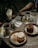 A wooden table is set with two plates of desserts, one with a chocolate cake and the other with a vanilla cake. Two cups of tea are placed on metal saucers, alongside sugar sticks. A small jar with paper napkins and another empty jar are also present. A printed paper with a QR code is partially visible on the table. The setting appears casual and warm, suggesting a cafe or outdoor dining environment.