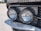 Close-up view of a vintage car's front headlights. The round headlights are encased in chrome and reflect light, with a grille visible in the background. The surroundings include some decorative coins attached to the vehicle near the headlights.