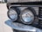 Close-up view of a vintage car's front headlights. The round headlights are encased in chrome and reflect light, with a grille visible in the background. The surroundings include some decorative coins attached to the vehicle near the headlights.