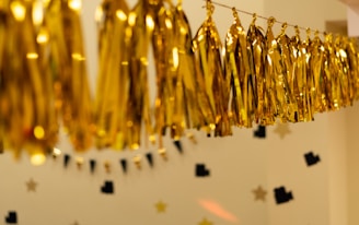 Golden metallic tassel garlands are hanging in a festive arrangement against a beige background. The wall is decorated with black and gold star-shaped paper cutouts, creating a celebratory atmosphere.