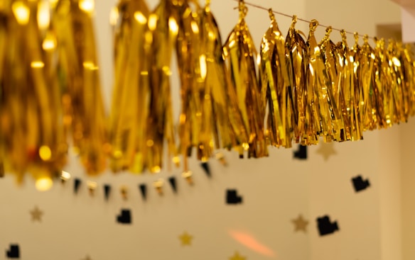 Golden metallic tassel garlands are hanging in a festive arrangement against a beige background. The wall is decorated with black and gold star-shaped paper cutouts, creating a celebratory atmosphere.