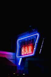 A neon sign illuminated with vibrant colors, specifically displaying the phrase 'Burger Bar'. The sign features a combination of blue and red neon lights against a dark background, which makes it stand out prominently. There is a partial view of a building with a tiled exterior and additional neon lights or signage.