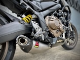 A close-up view of a motorcycle's exhaust system, showcasing the detailed design and engineering of the metallic parts. The image focuses on the Akrapovič exhaust muffler along with the motorcycle’s rear suspension, visible through a bright yellow coil spring. The background is a muted gray wall.