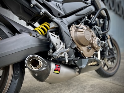 A close-up view of a motorcycle's exhaust system, showcasing the detailed design and engineering of the metallic parts. The image focuses on the Akrapovič exhaust muffler along with the motorcycle’s rear suspension, visible through a bright yellow coil spring. The background is a muted gray wall.
