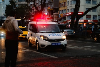 A police van with flashing red lights is driving through a busy city street. It is evening, as evident from the dark surroundings and artificial lighting. Several other vehicles, including a yellow taxi, are visible, along with a building featuring bright signage. There is a person in the foreground wearing a hooded sweatshirt, seemingly crossing the street.
