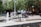 Children play in a fountain located in an urban plaza surrounded by trees and buildings. People are gathered around, some sitting and chatting under tents, and cyclists are seen nearby. The scene is lively and indicative of a community event or festival.