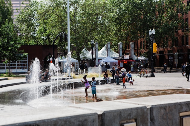 Children play in a fountain located in an urban plaza surrounded by trees and buildings. People are gathered around, some sitting and chatting under tents, and cyclists are seen nearby. The scene is lively and indicative of a community event or festival.