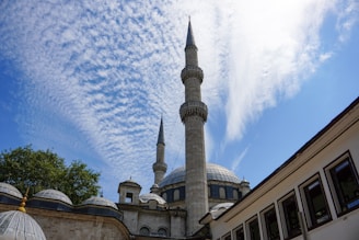 A mosque with two prominent minarets stands under a sky filled with scattered clouds. The building features a large central dome surrounded by several smaller domes. The architectural style is traditional and elegant, surrounded by trees on one side and a white building with rectangular windows on the other.