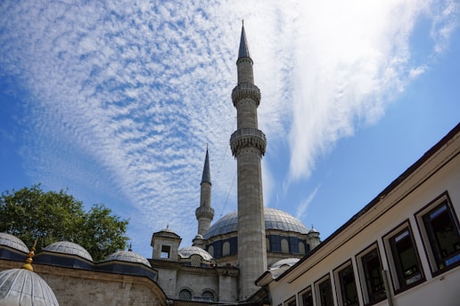 A mosque with two prominent minarets stands under a sky filled with scattered clouds. The building features a large central dome surrounded by several smaller domes. The architectural style is traditional and elegant, surrounded by trees on one side and a white building with rectangular windows on the other.