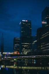 A night-time cityscape featuring a well-lit skyscraper with an HSBC logo prominently displayed at the top. The scene includes several tall buildings with lights in their windows reflecting off a body of water below. A bridge, also illuminated, spans across the water in the foreground.