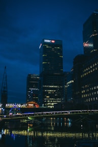 A night-time cityscape featuring a well-lit skyscraper with an HSBC logo prominently displayed at the top. The scene includes several tall buildings with lights in their windows reflecting off a body of water below. A bridge, also illuminated, spans across the water in the foreground.