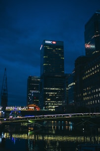A night-time cityscape featuring a well-lit skyscraper with an HSBC logo prominently displayed at the top. The scene includes several tall buildings with lights in their windows reflecting off a body of water below. A bridge, also illuminated, spans across the water in the foreground.