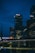 A night-time cityscape featuring a well-lit skyscraper with an HSBC logo prominently displayed at the top. The scene includes several tall buildings with lights in their windows reflecting off a body of water below. A bridge, also illuminated, spans across the water in the foreground.
